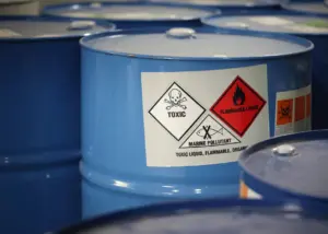 Blue industrial drums with hazard warning labels indicating toxic, flammable liquid, and marine pollutant contents, arranged in a storage facility.