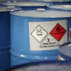 Blue industrial drums with hazard warning labels indicating toxic, flammable liquid, and marine pollutant contents, arranged in a storage facility.