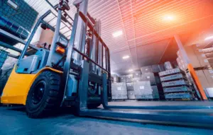 A yellow and blue forklift with an overhead hoist system in a modern warehouse interior, surrounded by stacked cardboard boxes on shelves and storage racks, illuminated by industrial lighting from a corrugated metal ceiling.