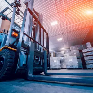 A yellow and blue forklift with an overhead hoist system in a modern warehouse interior, surrounded by stacked cardboard boxes on shelves and storage racks, illuminated by industrial lighting from a corrugated metal ceiling.