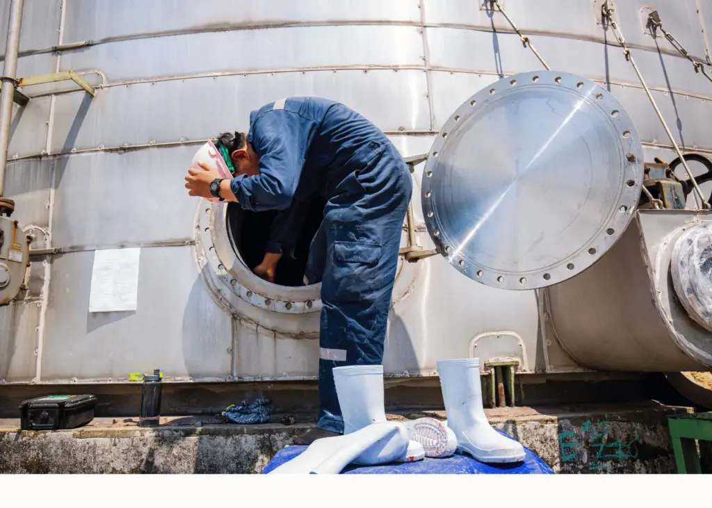 A worker in blue coveralls and safety glasses bends forward to inspect the interior of a large industrial storage tank, with the circular hatch door open beside them and protective equipment visible at ground level.