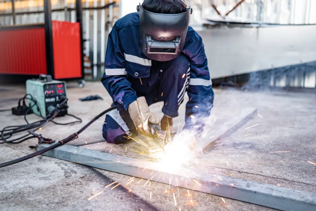 A welder wearing protective gear and a dark welding helmet performs hot work on a metal plate in an industrial outdoor setting, with sparks flying from the welding torch and welding equipment visible in the background.
