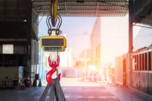 Close-up view of an overhead gantry crane hook with a yellow digital weight display showing "2" and a red hook mechanism suspended from steel cables inside an industrial warehouse facility with metal storage containers and bright sunlight streaming through the open bay doors.