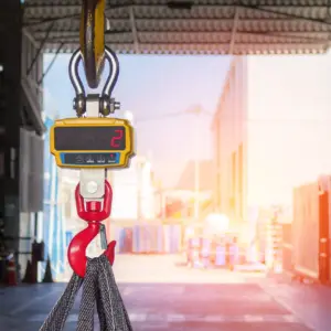 Close-up view of an overhead gantry crane hook with a yellow digital weight display showing "2" and a red hook mechanism suspended from steel cables inside an industrial warehouse facility with metal storage containers and bright sunlight streaming through the open bay doors.