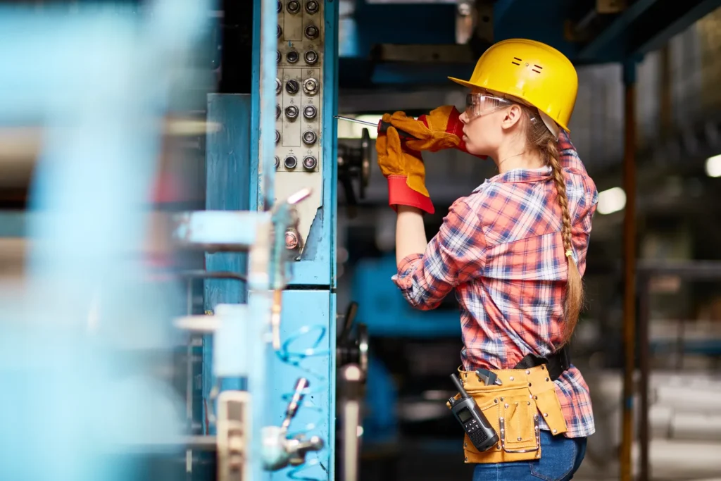Construction worker wearing yellow hard hat, safety glasses, and work gloves inspecting electrical wiring and connections on blue metal panel with multiple circuit breakers and switches in an industrial workshop setting.