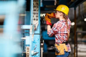 Construction worker wearing yellow hard hat, safety glasses, and work gloves inspecting electrical wiring and connections on blue metal panel with multiple circuit breakers and switches in an industrial workshop setting.