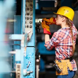 Construction worker wearing yellow hard hat, safety glasses, and work gloves inspecting electrical wiring and connections on blue metal panel with multiple circuit breakers and switches in an industrial workshop setting.