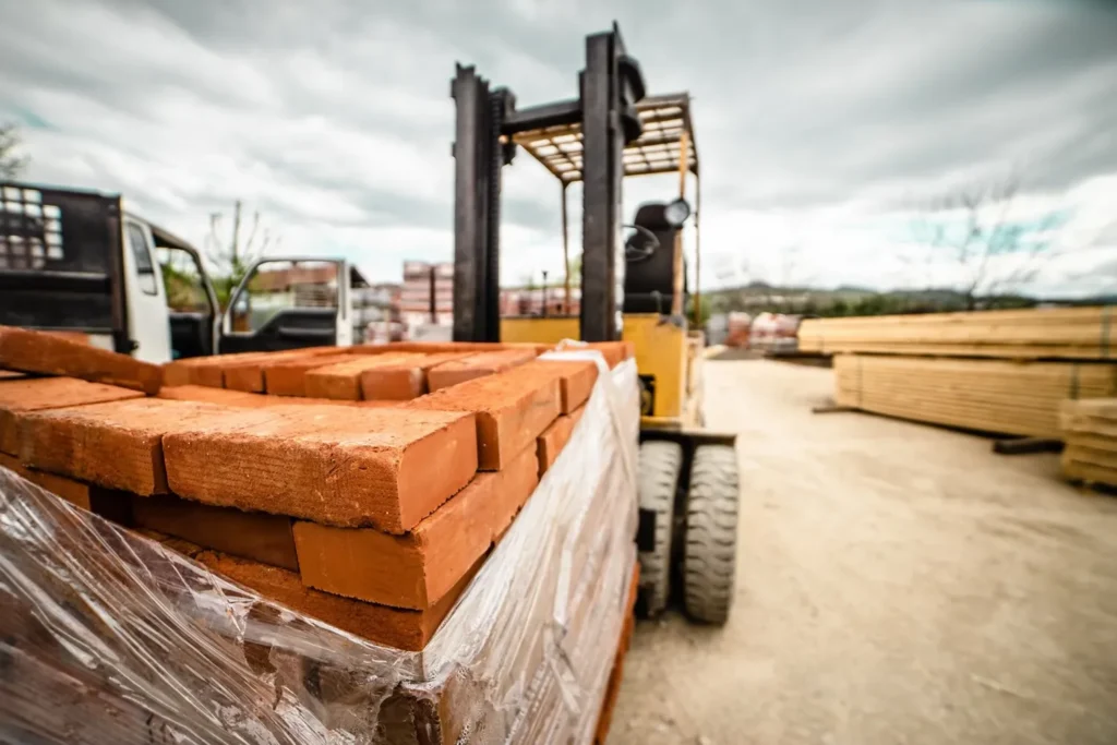 Yellow forklift loading red bricks onto a pallet in an outdoor construction yard with wooden lumber and building materials visible in the background under a cloudy sky.
