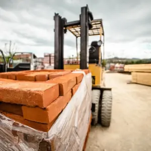 Yellow forklift loading red bricks onto a pallet in an outdoor construction yard with wooden lumber and building materials visible in the background under a cloudy sky.