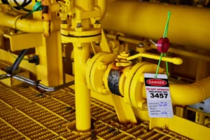 Bright yellow industrial pipeline and valve equipment with a red and white danger tag labeled "DO NOT OPERATE ISOLATION TAG 3457" attached with a green wire lock, indicating equipment under lockout-tagout safety protocol.
