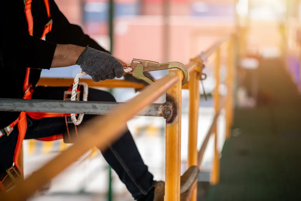 A construction worker wearing a red and black safety harness and gray work gloves secures a carabiner clip to a wooden railing at height, demonstrating proper fall protection equipment and procedures on a job site.