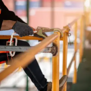 A construction worker wearing a red and black safety harness and gray work gloves secures a carabiner clip to a wooden railing at height, demonstrating proper fall protection equipment and procedures on a job site.