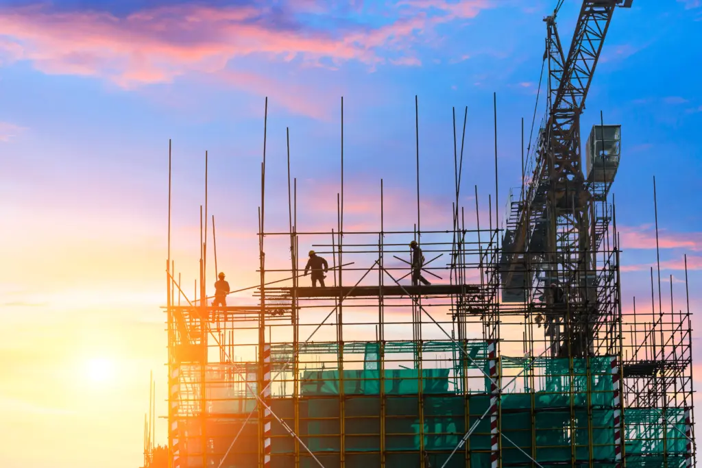 Construction workers silhouetted against a colorful sunset sky work on a large metal scaffold structure with a crane visible on the right side of the frame.
