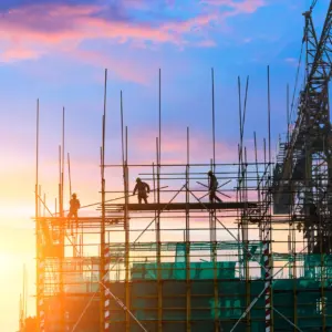 Construction workers silhouetted against a colorful sunset sky work on a large metal scaffold structure with a crane visible on the right side of the frame.